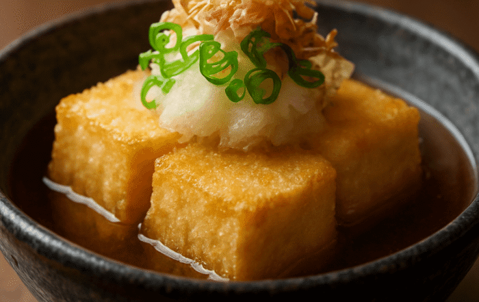 Warm Japanese agedashi tofu in dashi broth, topped with daikon, green onion, and bonito flakes, in a black bowl with rising steam.