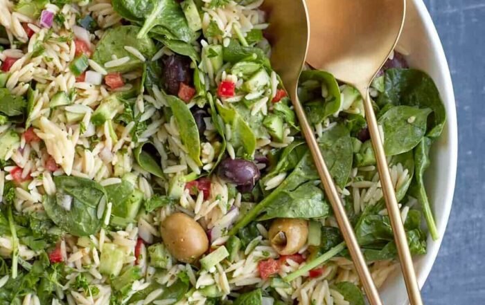Greek orzo pasta salad with spinach, cucumber, peppers, olives, and gold serving spoons, in a white bowl on a dark blue background.