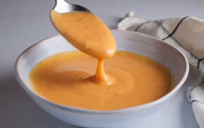 Creamy orange Bang Bang Sauce being lifted with a spoon over a gray bowl, with a checkered cloth in the background.