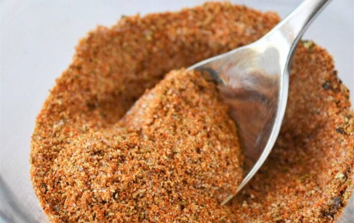 Coarse reddish-orange French fry seasoning in a glass bowl with a metal spoon, on a marble surface.