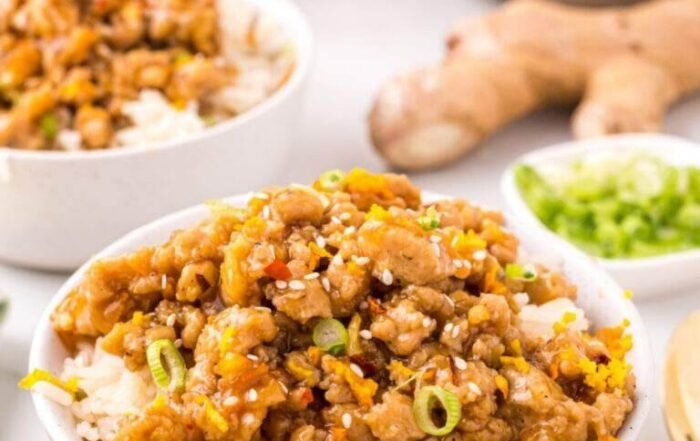 Garnished ground orange chicken over rice in a white bowl, with ginger, scallions, and an orange slice in the background.