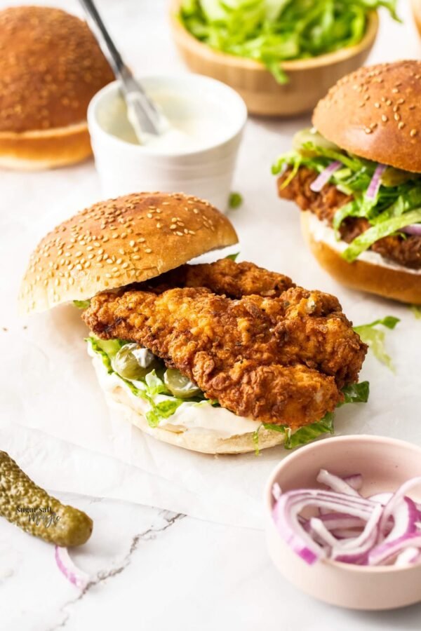 Partially open crispy chicken burger with patty and toppings, plus a second burger and topping bowls, on a light table.