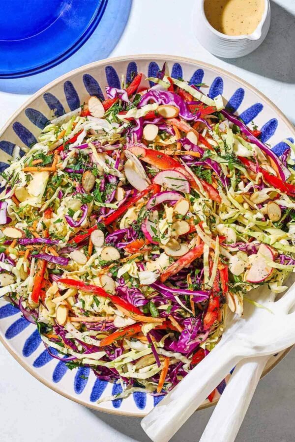Crunchy green and purple cabbage coleslaw with carrots, tossed in lemon dressing, in a glass bowl with a spoon, lemon wedges, and pepper nearby.
