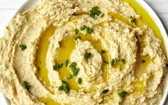 Creamy hummus with olive oil drizzle and chives, served with a lemon half and whole-grain crackers on a white surface.