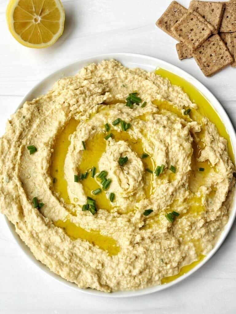 Creamy hummus with olive oil drizzle and chives, served with a lemon half and whole-grain crackers on a white surface.