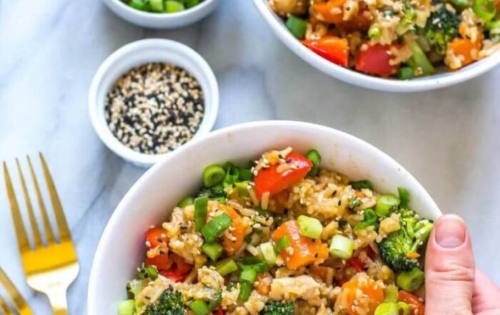 Teriyaki chicken fried rice bowls (with broccoli, carrots, peppers) topped with scallions/sesame seeds, held by a hand, with garnishes and gold forks in the background.