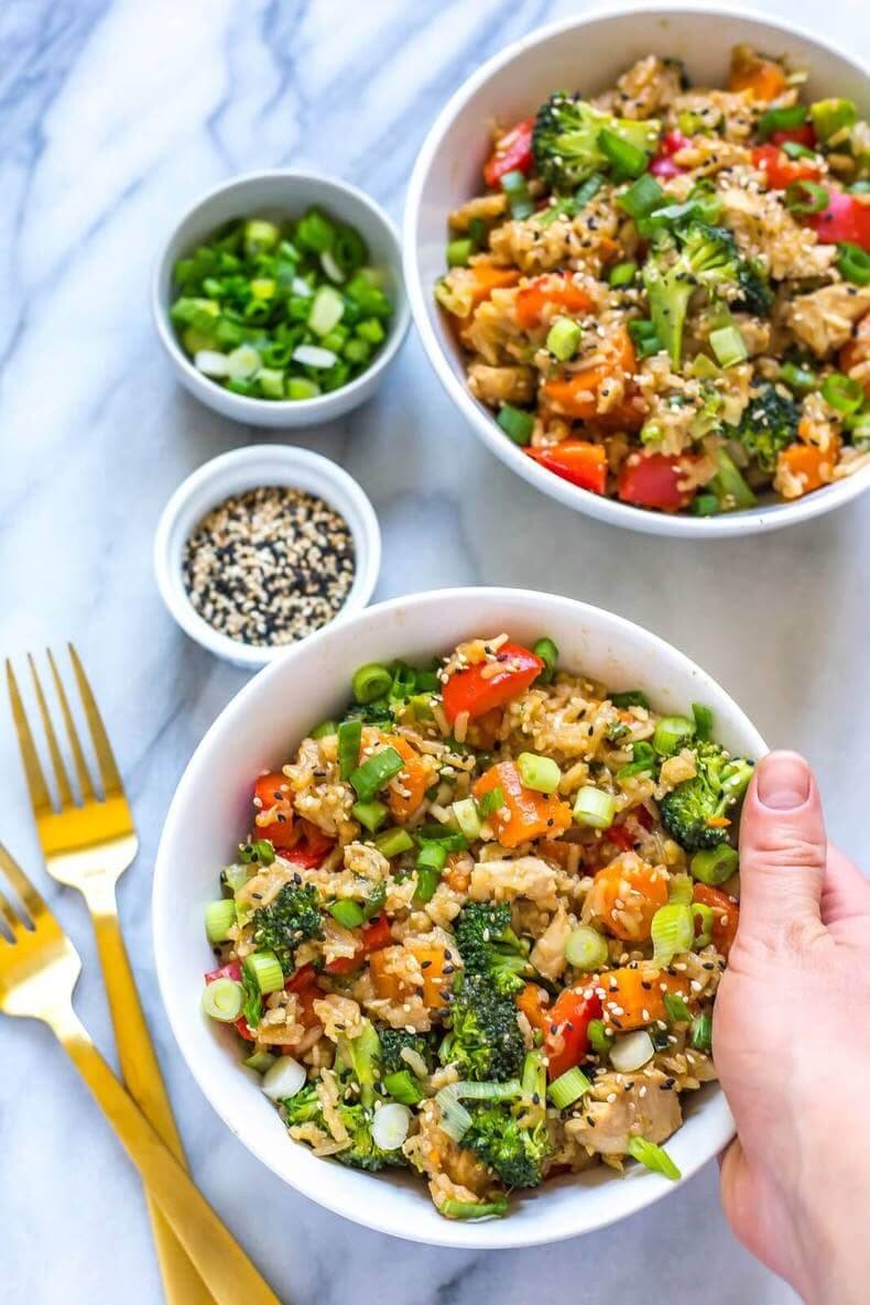Teriyaki chicken fried rice bowls (with broccoli, carrots, peppers) topped with scallions/sesame seeds, held by a hand, with garnishes and gold forks in the background.
