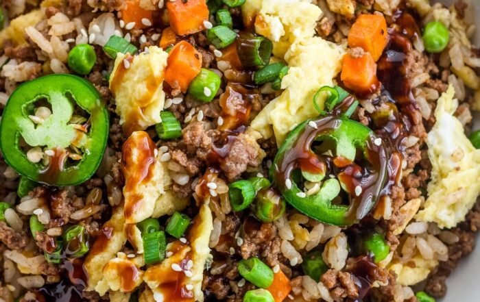 Loaded ground beef fried rice in a white bowl, topped with jalapeños, scallions, sesame seeds, and sauce, with extra jalapeños in the background.