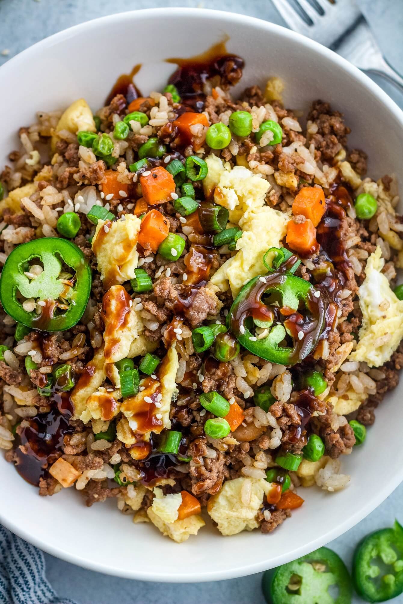 Loaded ground beef fried rice in a white bowl, topped with jalapeños, scallions, sesame seeds, and sauce, with extra jalapeños in the background.