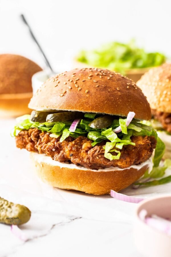 Crispy chicken burger with lettuce, pickles, red onion, and sauce, in a sesame seed bun on a marble surface.