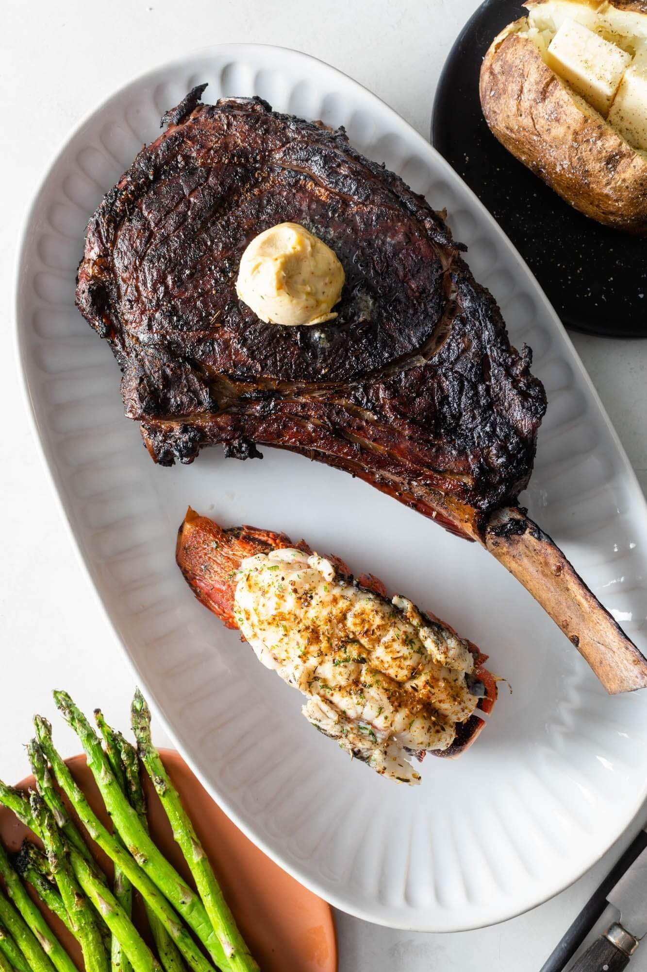 Char-grilled tomahawk steak with compound butter, stuffed lobster tail, grilled asparagus, and a baked potato, on a white platter.